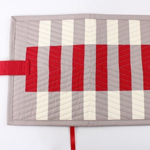May include: A grey and white quilted fabric pouch with a red strap and a red and white striped fabric panel. The pouch has a ribbon tie closure.