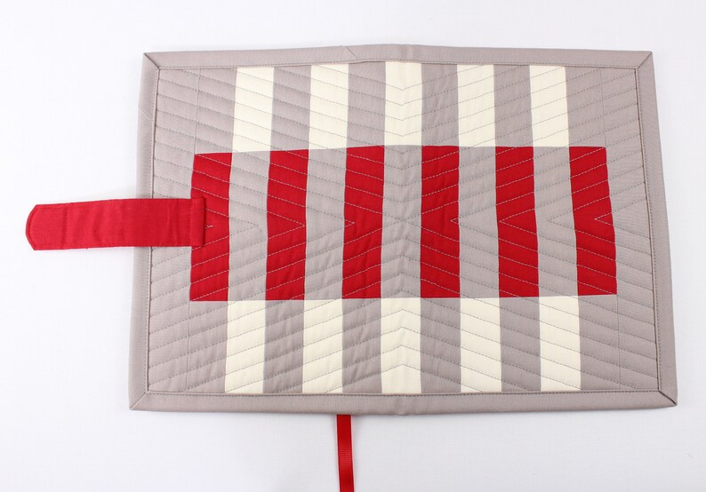 May include: A grey and white quilted fabric pouch with a red strap and a red and white striped fabric panel. The pouch has a ribbon tie closure.