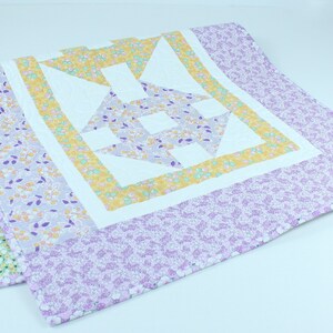 Puede incluir: Una colcha blanca con un patr&oacute;n floral morado y amarillo. La colcha tiene un gran dise&ntilde;o geom&eacute;trico en el centro.