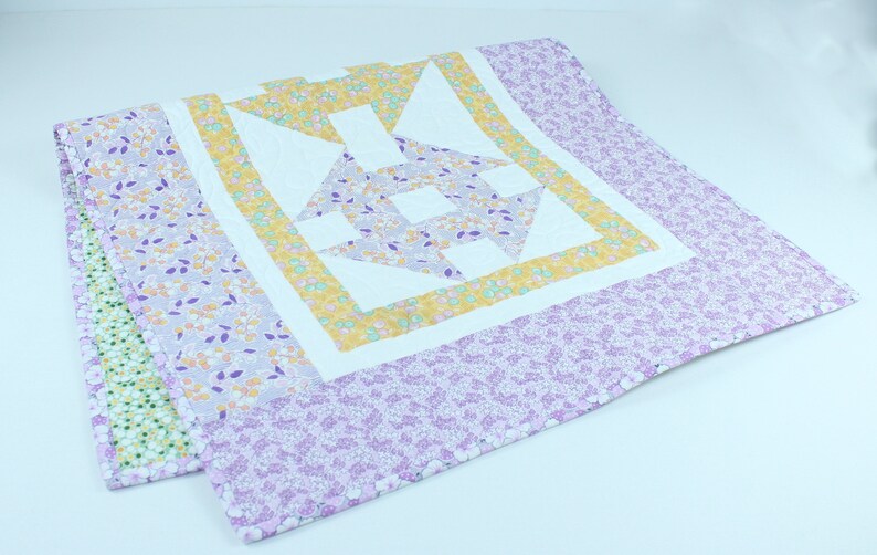 Puede incluir: Una colcha blanca con un patr&oacute;n floral morado y amarillo. La colcha tiene un gran dise&ntilde;o geom&eacute;trico en el centro.
