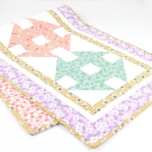 May include: A white quilted table runner with a patchwork design featuring pink, green, and yellow floral patterns. The runner has a white background with a yellow, pink, and purple floral border.