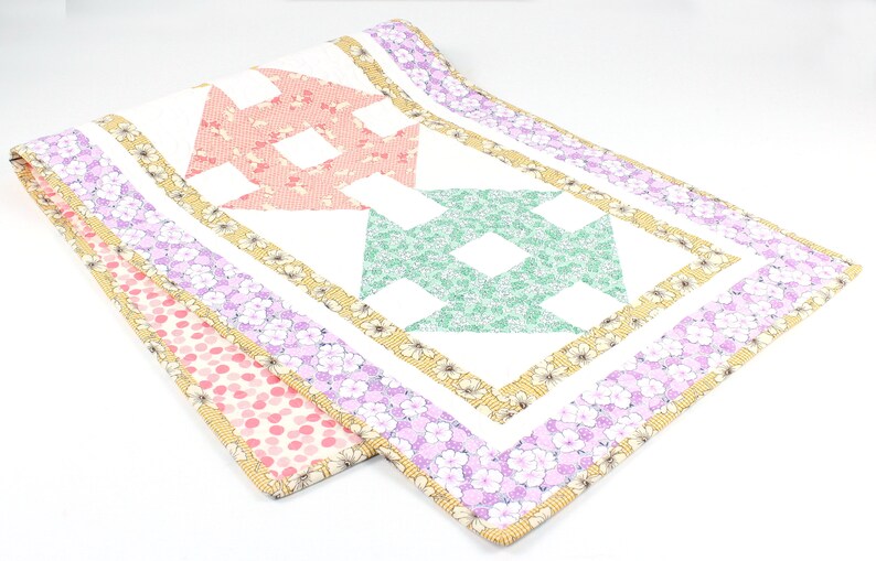 May include: A white quilted table runner with a patchwork design featuring pink, green, and yellow floral patterns. The runner has a white background with a yellow, pink, and purple floral border.