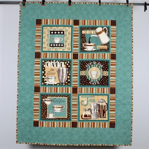 May include: A teal blue quilted fabric wall hanging with a brown and white striped border. The center of the wall hanging features six squares with coffee-themed designs. Each square has a different design, including coffee cups, coffee pots, and coffee beans. The text on the squares includes "Cafe de Jour", "Coffee", "Java", "Cup o Joe", "Coffee Classico", and "Good Coffee".