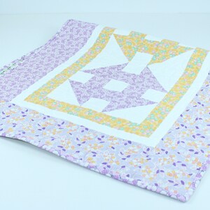 Puede incluir: Una colcha de retazos de color morado, amarillo y blanco con un patr&oacute;n floral. La colcha est&aacute; doblada por la mitad y se encuentra sobre una superficie blanca.