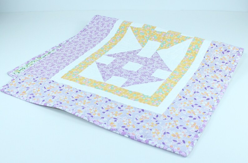 Puede incluir: Una colcha de retazos de color morado, amarillo y blanco con un patr&oacute;n floral. La colcha est&aacute; doblada por la mitad y se encuentra sobre una superficie blanca.