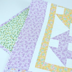 Puede incluir: Una colcha de retazos de color morado, amarillo y verde con un patr&oacute;n geom&eacute;trico. La colcha est&aacute; hecha de diferentes telas con estampados florales.