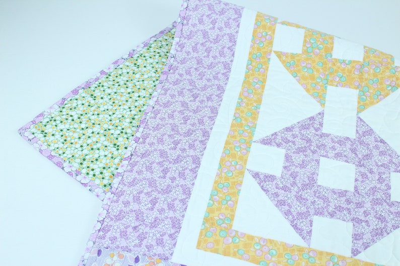 Puede incluir: Una colcha de retazos de color morado, amarillo y verde con un patr&oacute;n geom&eacute;trico. La colcha est&aacute; hecha de diferentes telas con estampados florales.