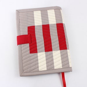May include: A grey and white striped fabric book cover with a red and white gingham pattern on the front. The cover has a red fabric strap and a red fabric bookmark.