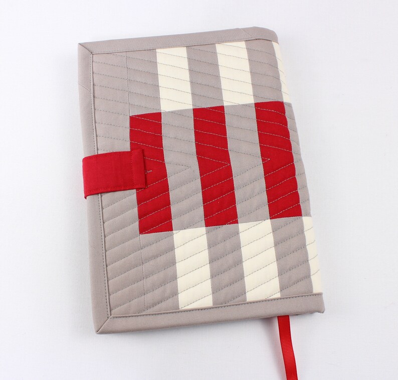 May include: A grey and white striped fabric book cover with a red and white gingham pattern on the front. The cover has a red fabric strap and a red fabric bookmark.
