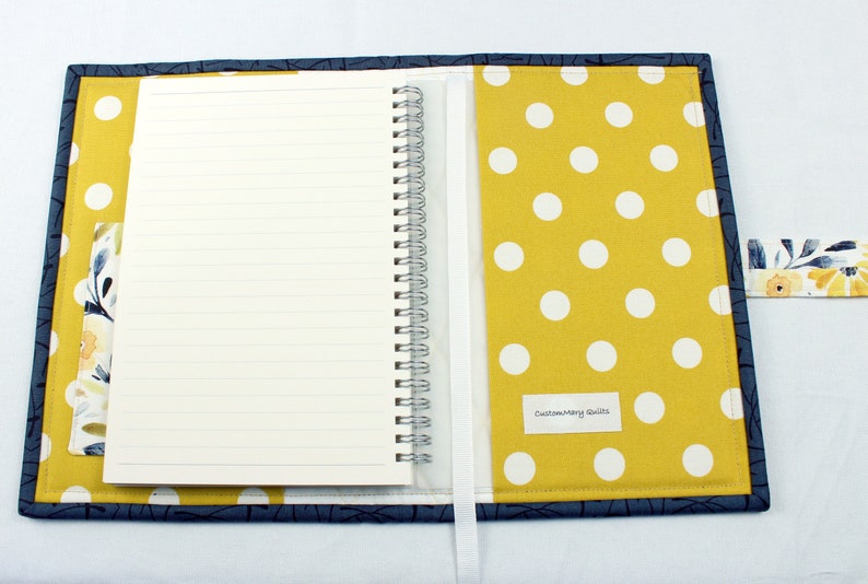 May include: A yellow and white polka dot fabric notebook cover with a blue floral fabric pocket and a white ribbon bookmark. The cover has a blue fabric border and the text "Custom Mary Quilts" is printed on the front.