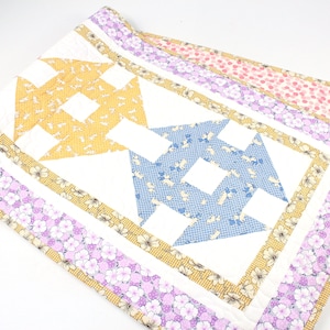 May include: A white quilted table runner with a geometric pattern of yellow, blue, and white fabric squares. The runner has a border of yellow, white, and purple floral fabric.