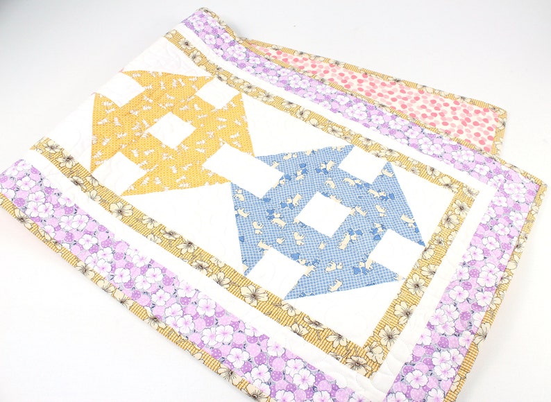 May include: A white quilted table runner with a geometric pattern of yellow, blue, and white fabric squares. The runner has a border of yellow, white, and purple floral fabric.