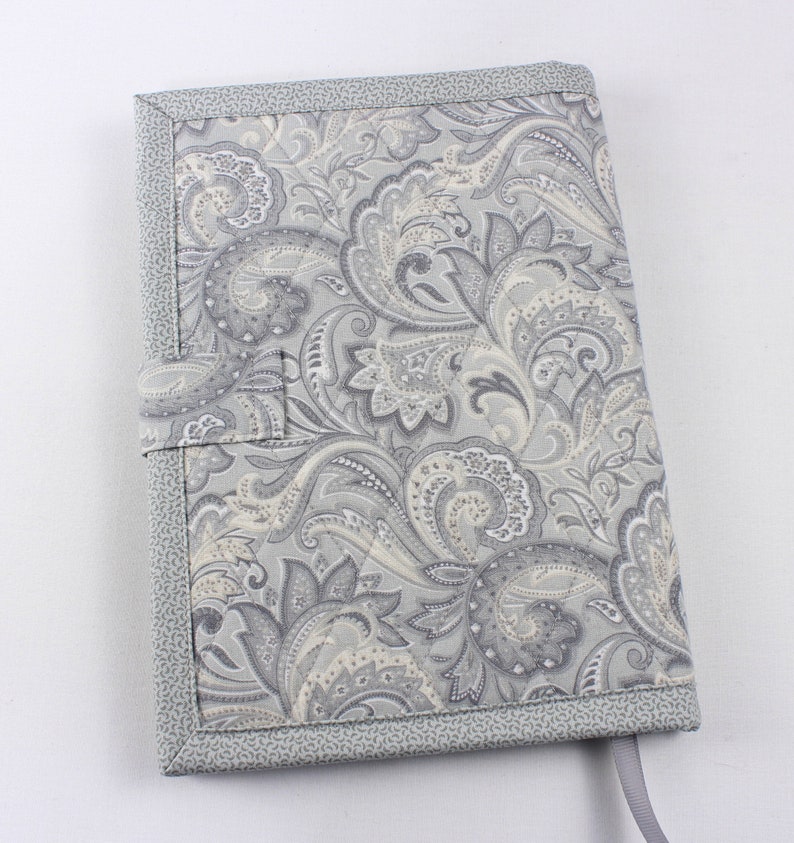 Pu&ograve; includere: Una copertina per libro con motivo paisley grigio e bianco con un bordo in tessuto grigio e un nastro grigio.
