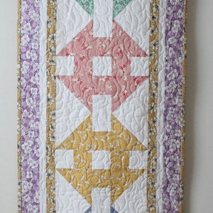 May include: A quilted wall hanging with a white background and a pattern of five colourful squares. The squares are in shades of green, pink, yellow, blue, and purple. The squares are surrounded by a white border and a purple and yellow floral border.
