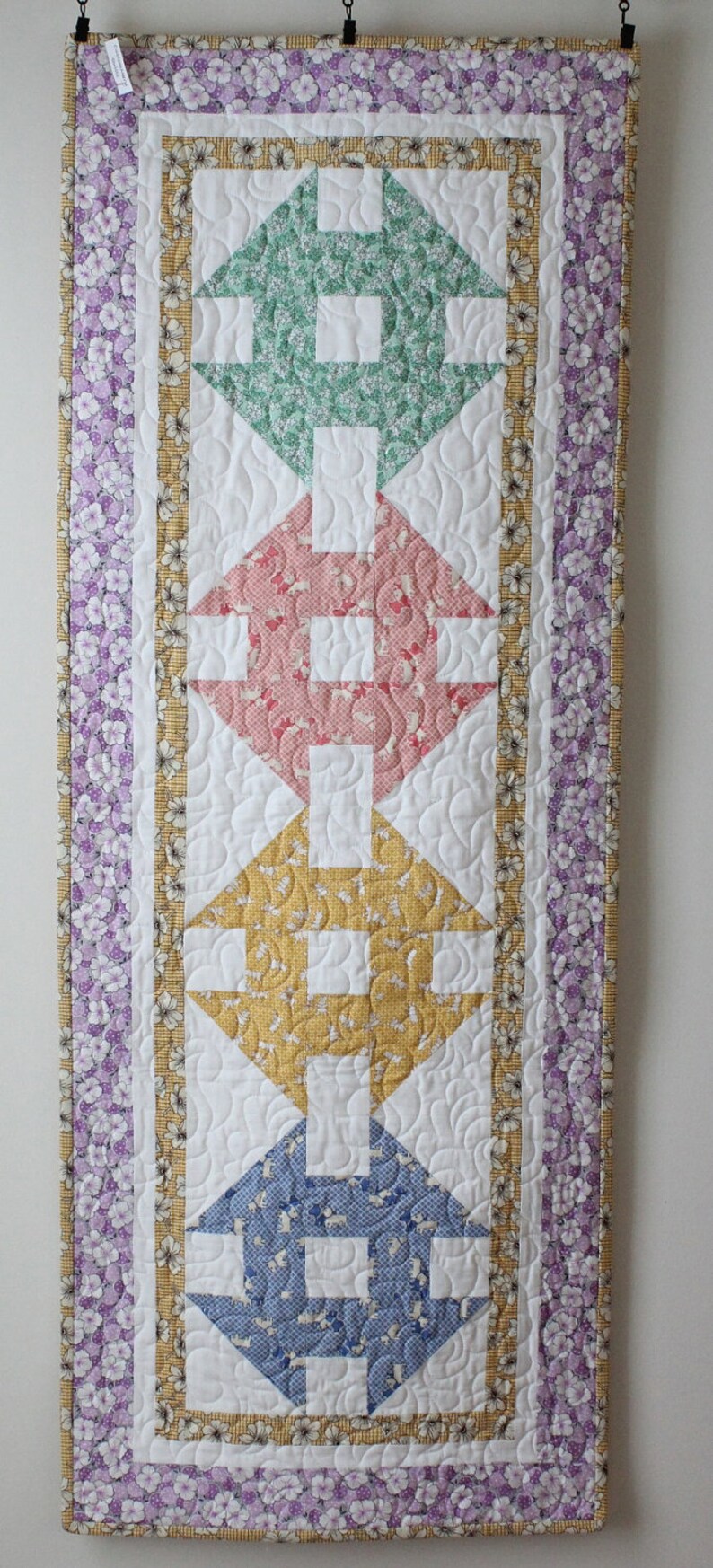 May include: A quilted wall hanging with a white background and a pattern of five colourful squares. The squares are in shades of green, pink, yellow, blue, and purple. The squares are surrounded by a white border and a purple and yellow floral border.