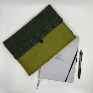 May include: A two-tone green felt notebook sleeve with a black closure, a silver notebook with a black elastic band, and a black pen. The sleeve is olive green and dark green.