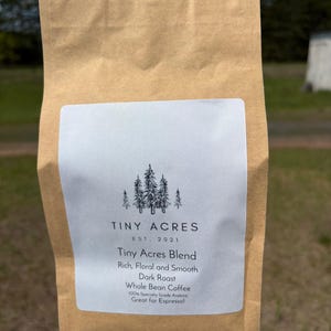 May include: A brown paper bag of Tiny Acres Blend whole bean coffee. The label reads "Rich, Floral and Smooth Dark Roast." The bag is sealed and ready for purchase. The background is a blurred outdoor scene.