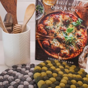 May include: A white ceramic utensil holder containing wooden spoons and a honey dipper, a cookbook titled Half Baked Harvest Quick & Cozy by Tieghan Gerard, and two round felt ball trivets in shades of grey and olive green.