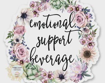 May include: A sticker with a floral wreath design. The wreath is composed of pink, purple, and green flowers. The text "emotional support beverage" is written in black script in the center. The sticker has a white background.