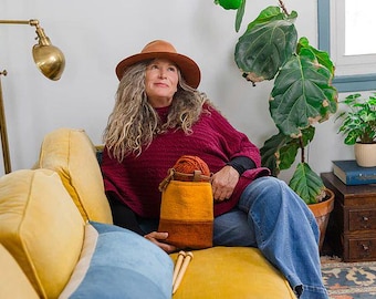 May include: A woman wearing a brown hat and a burgundy poncho sits on a yellow couch, holding an orange and brown yarn bag. The bag contains a ball of orange yarn. A brass lamp and potted plants are in the background.