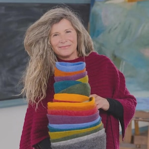 May include: A woman holding a stack of colorful felted wool bowls. The bowls are in various colors, including blue, green, orange, and purple. The woman is wearing a burgundy sweater. The background is a blurred painting.