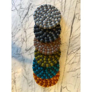 May include: A stack of seven round felt trivets in various colors, including gray, navy, brown, olive green, teal, and orange. Each trivet is made of small, tightly packed felt balls. The trivets are arranged vertically on a marble surface.