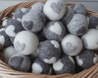 May include: A wicker basket filled with gray and white felt balls. Each ball has a heart design. The balls are a mix of solid gray and white, with the heart design in the opposite color. The basket is made of woven brown wicker.