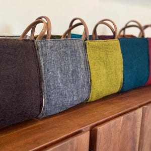 Op de afbeelding: Zes gevoerde tassen met leren handvatten. De tassen zijn in een rij op een houten oppervlak gerangschikt. De tassen zijn in verschillende kleuren, waaronder bruin, grijs, geel, teal, rood en oranje.