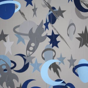 May include: A blue and gray paper mobile with rockets, planets, stars, and moons. The mobile is made of paper and is perfect for a baby's nursery.