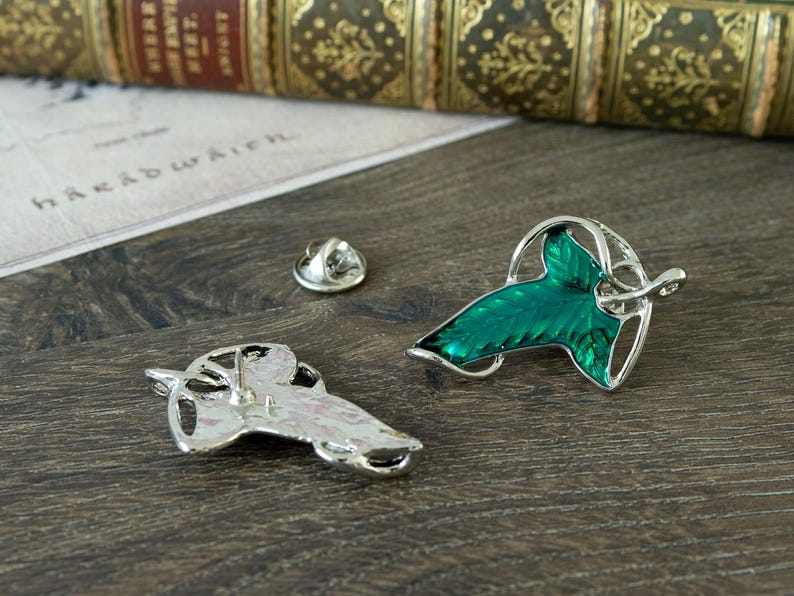 May include: Two silver-toned metal pins shaped like leaves with vibrant green enamel filling. One pin is displayed front-facing showing the green leaf detail, while the other is flipped to show the silver back and pin post. A small circular metal clutch is included.