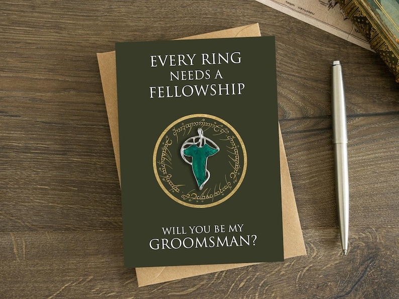 May include: A dark olive green greeting card featuring the text EVERY RING NEEDS A FELLOWSHIP and WILL YOU BE MY GROOMSMAN? with a central graphic of a gold circular ring surrounding a silver and green leaf-shaped brooch pin.