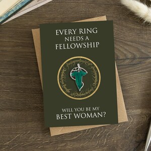 May include: A dark olive green greeting card featuring the text EVERY RING NEEDS A FELLOWSHIP and WILL YOU BE MY BEST WOMAN? The center displays a circular gold emblem with script surrounding a silver and green leaf-shaped brooch pin.