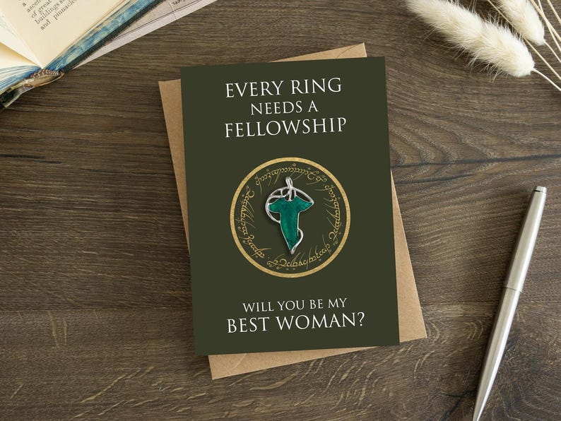May include: A dark olive green greeting card featuring the text EVERY RING NEEDS A FELLOWSHIP and WILL YOU BE MY BEST WOMAN? The center displays a circular gold emblem with script surrounding a silver and green leaf-shaped brooch pin.