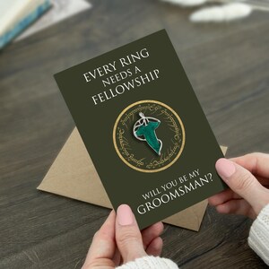 May include: A dark olive green greeting card featuring the text "EVERY RING NEEDS A FELLOWSHIP" and "WILL YOU BE MY GROOMSMAN?" with a central circular graphic containing a silver and green leaf-shaped brooch design on a gold ring background.