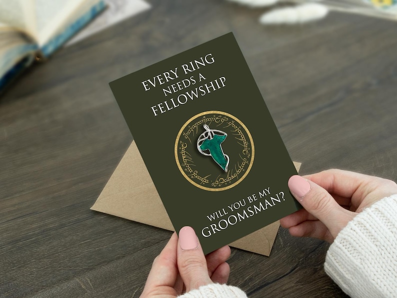 May include: A dark olive green greeting card featuring the text "EVERY RING NEEDS A FELLOWSHIP" and "WILL YOU BE MY GROOMSMAN?" with a central circular graphic containing a silver and green leaf-shaped brooch design on a gold ring background.