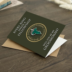 May include: A dark olive green greeting card featuring the text EVERY RING NEEDS A FELLOWSHIP and WILL YOU BE MY GROOMSMAN? The card displays a circular gold emblem with script and a silver-toned leaf-shaped brooch with green enamel detailing.