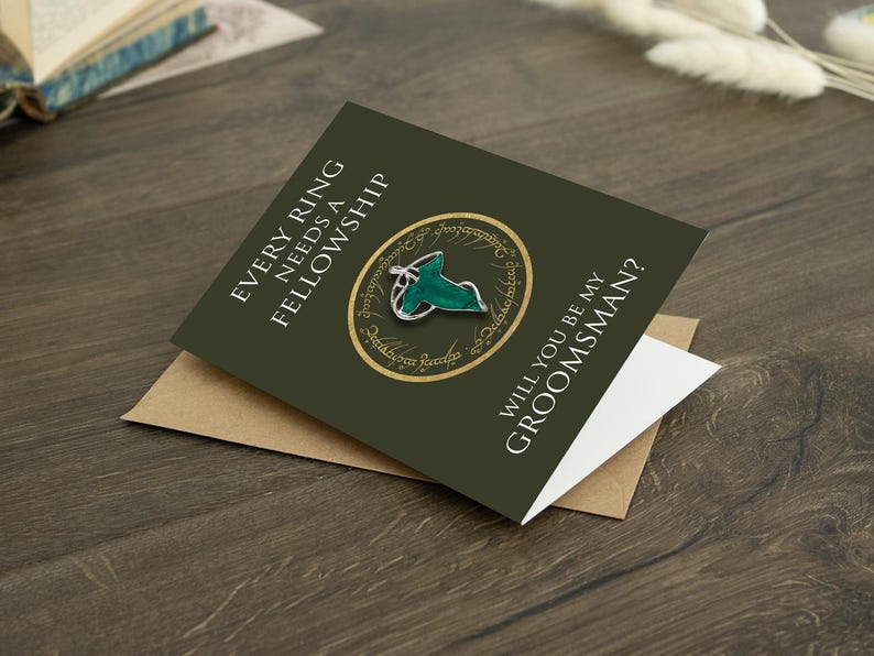 May include: A dark olive green greeting card featuring the text EVERY RING NEEDS A FELLOWSHIP and WILL YOU BE MY GROOMSMAN? The card displays a circular gold emblem with script and a silver-toned leaf-shaped brooch with green enamel detailing.