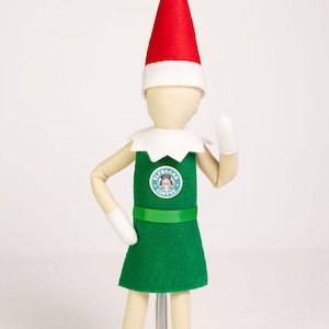 May include: A doll wearing a red pointed hat with a white brim, a white collar, and a green apron over a light-colored body. The apron features a circular logo with the text Elfbucks Coffee. The doll has white mittens and articulated limbs.