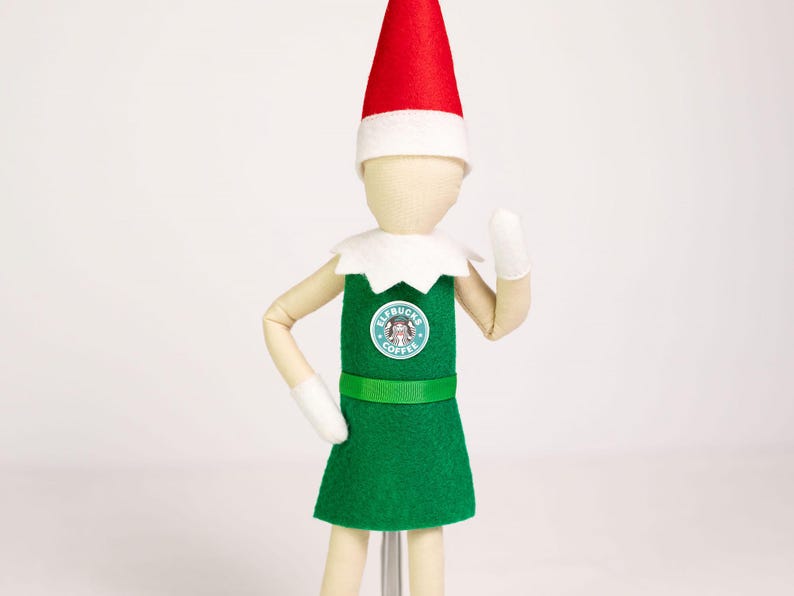 May include: A doll wearing a red pointed hat with a white brim, a white collar, and a green apron over a light-colored body. The apron features a circular logo with the text Elfbucks Coffee. The doll has white mittens and articulated limbs.
