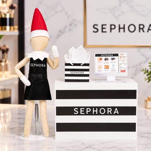 May include: A doll wearing a red and white hat and a black apron with the Sephora logo, standing next to a white counter with black stripes and the Sephora logo. On the counter sits a small black and white striped shopping bag and a computer monitor.