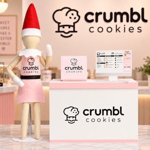 May include: A miniature Crumbl Cookies shop display featuring a doll wearing a red hat and a pink apron with the Crumbl Cookies logo. The setup includes a white counter with the logo, a small digital menu screen, and a sign with the Crumbl Cookies logo.