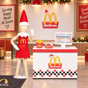 May include: A beige doll wearing a red felt uniform and hat stands beside a white service counter with a red and black checkered base. The counter features a McDonald's logo with a Santa hat, a cash register, fries, and a burger box.