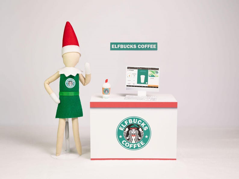 May include: A doll wearing a red pointed hat, white collar, and green apron with an ELFBUCKS COFFEE logo stands beside a white counter. The counter features an ELFBUCKS COFFEE logo, a small coffee cup, and a computer monitor displaying a menu.