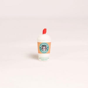 May include: A miniature white coffee cup with a tan sleeve featuring a green circular logo that reads ELFBUCKS COFFEE and a graphic of an elf. The cup is topped with white foam and a small red straw. Text at the bottom reads: Careful, the beverage you're about to enjoy is very hot.
