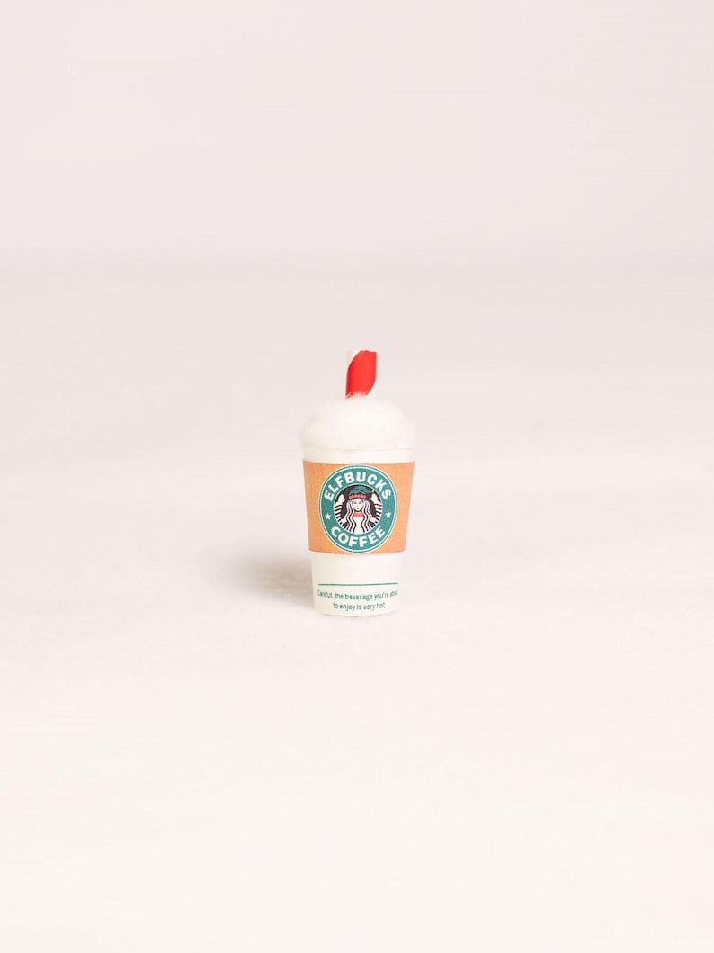 May include: A miniature white coffee cup with a tan sleeve featuring a green circular logo that reads ELFBUCKS COFFEE and a graphic of an elf. The cup is topped with white foam and a small red straw. Text at the bottom reads: Careful, the beverage you're about to enjoy is very hot.