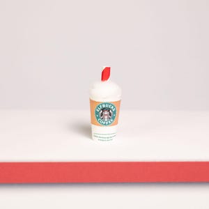 May include: A miniature white coffee cup with a red and white striped straw. The cup features a tan sleeve with a circular green logo containing a graphic of an elf and the text ELFBUCKS COFFEE. The top of the cup is filled with white foam.