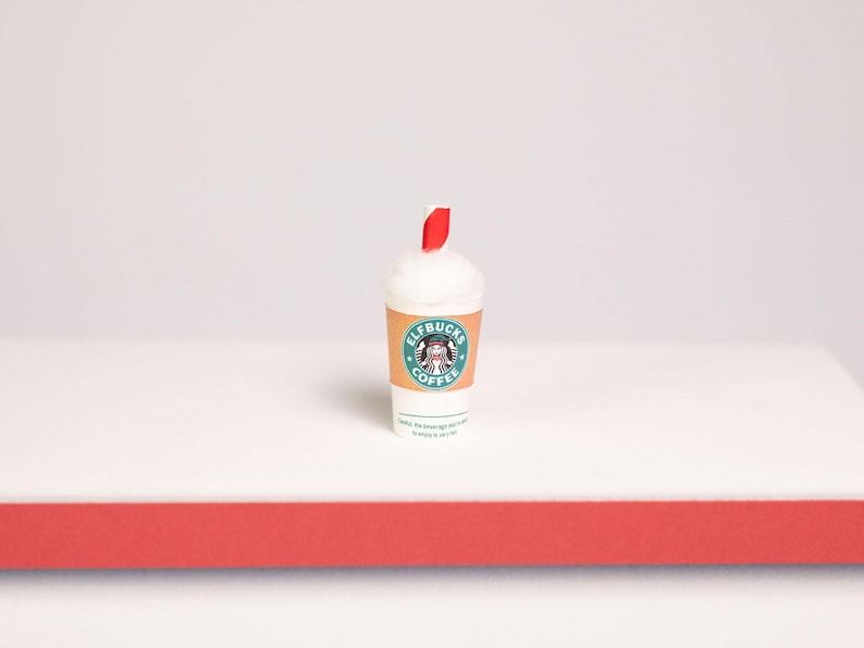 May include: A miniature white coffee cup with a red and white striped straw. The cup features a tan sleeve with a circular green logo containing a graphic of an elf and the text ELFBUCKS COFFEE. The top of the cup is filled with white foam.