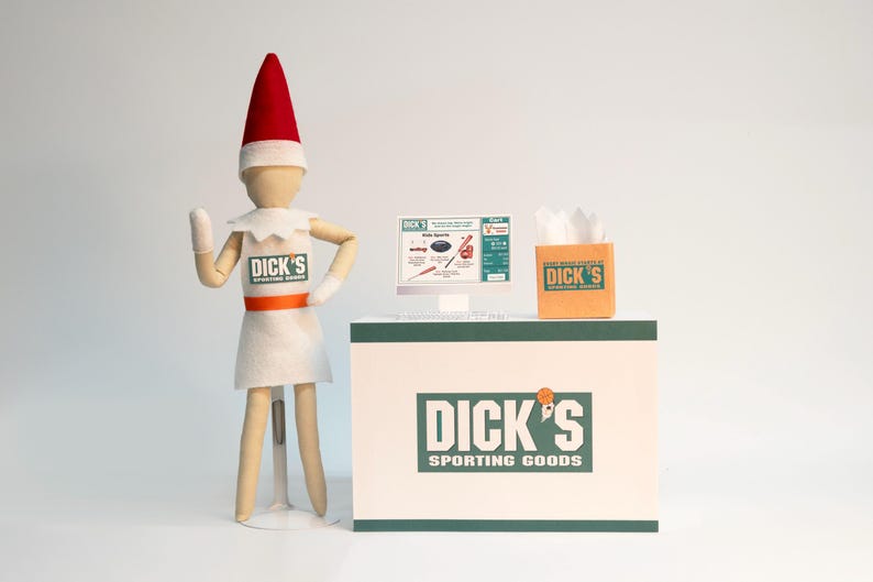 May include: A miniature elf figurine wearing a white apron with the Dick's Sporting Goods logo, standing beside a checkout counter. The counter features a small computer screen displaying a shopping cart and a brown paper bag with the store logo.