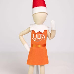 Puede incluir: Mu&ntilde;eca de fieltro con forma de elfo, vestida con un vestido naranja con el logo "ULTA BEAUTY", un cuello blanco, guantes blancos y un gorro rojo y blanco. La mu&ntilde;eca est&aacute; sobre un fondo blanco.