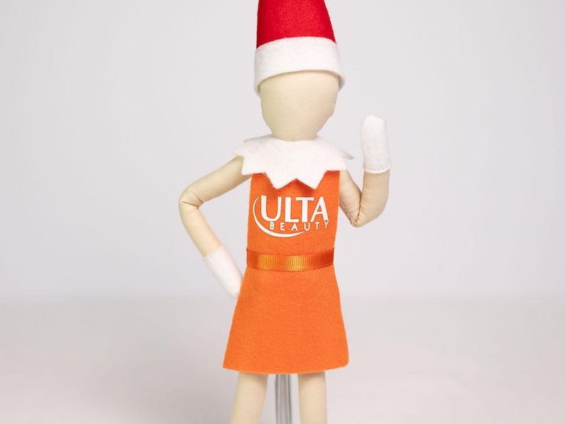 Puede incluir: Mu&ntilde;eca de fieltro con forma de elfo, vestida con un vestido naranja con el logo "ULTA BEAUTY", un cuello blanco, guantes blancos y un gorro rojo y blanco. La mu&ntilde;eca est&aacute; sobre un fondo blanco.